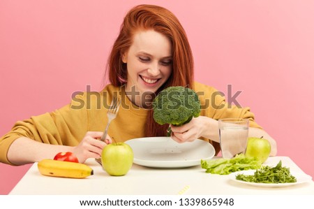 Hungry young vegg woman eating with eagerness and appetite raw green broccoli , smiling happy at camera over studio isolated pink background. Vegetable diet.
