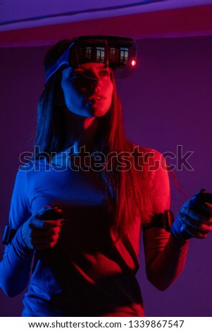Young woman with long ginger hair sets programm for VR device to se 3 D movie, holding handheld controllers over dark background with red, blue , purple color