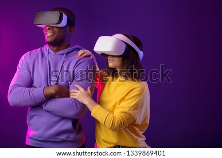 Technology and innovation concept. Two multicultural cheerful people, dressed in yellow and violet coloures, sitting side by side with VR goggles over purple background, having 3d tour