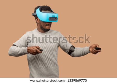Dark skinned male gamer wearing virtual reality goggles, playing 3d game, isolated against studio beige wall.