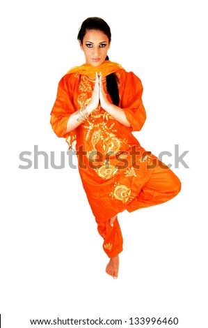 A beautiful young east indian teen girl standing on one foot in her nice traditional indian orange dress, folding her hands for white background.