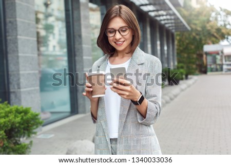 Young businesswoman wearing eyeglasses standing on the city street holding cup of hot coffee browsing internet on smartphone smiling happy