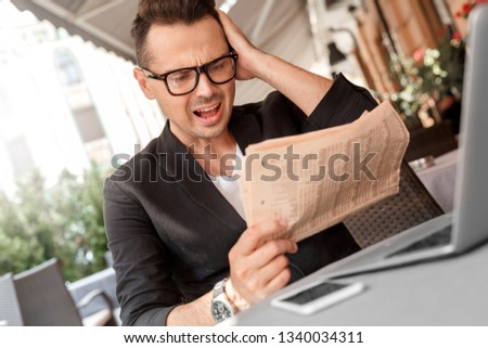 Young stylish man wearing suit and eyeglasses sitting at the table in cafe on the city street with laptop holding newspaper reading stock news touching head concerned close-up.