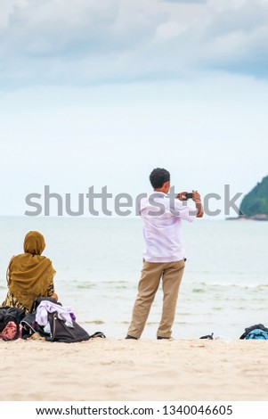Rear view of a Muslims family relaxing on the sand beach and taking photo with smartphone of a tropical island. Samila, Thailand.