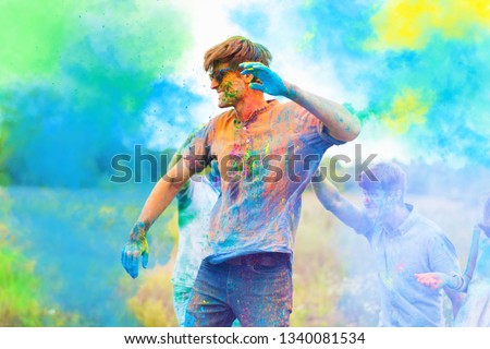 Happy positive smiling fun man wearing blue sunglasses all stained with colorful paint celebrating Holi festival party with friends. On color dust smoke powder cloud background in park field outdoor.  Royalty-Free Stock Photo #1340081534
