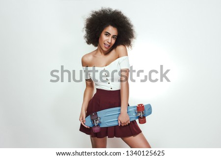 I love skating! Studio shot of beautiful young African woman in casual wear holding skateboard and smiling while standing against grey background. Active lifestyle.