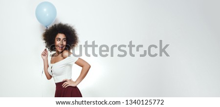 It's my party! Happy and beautiful afro American woman is holding blue balloon, keeping arm on hip and looking away with opened mouth. Space for text. Web Banner