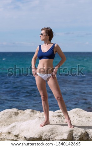 Portrait of barefoot thirty-nine-year-old woman at sea. Female person in sunglasses, sport bra and white panties posing with hands on hips on beach. She stands on white coastal rock and looking away