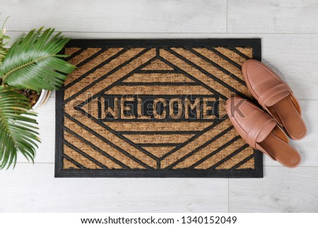 Door mat with word WELCOME and shoes on floor, top view