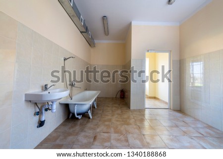Interior of spacious light hospital or kindergarten bathroom with white bathtub, sink, air ventilation duct and tiled floor and walls.