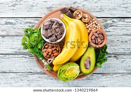 Foods containing natural magnesium. Mg: Chocolate, banana, cocoa, nuts, avocados, broccoli, almonds. Top view. On a white wooden background. Royalty-Free Stock Photo #1340219864
