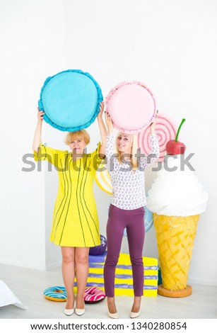 Two blonde women holding big cakes and posing on decorated background. Amazing sweet-tooth women surrounded by toy sweets. Vertical portrait
