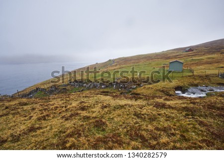 Landscape Picture of countryside in Faroe islands shown sheep grazing or pasture land in moorland on top of hill in fjord, wired fences, wild creek with water cascade during typical spring rainy day.