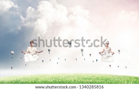 Young couple keeping eyes closed and looking concentrated while meditating on clouds among flying aerostats with bright and beautiful landscape on background.