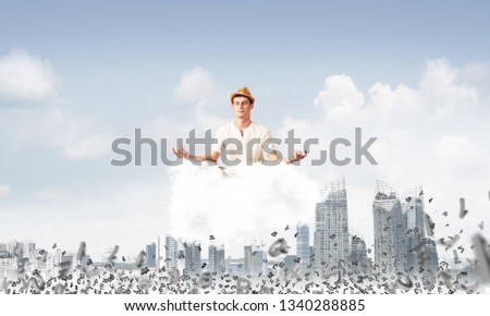 Man in white clothing keeping eyes closed and looking concentrated while meditating on cloud among flying letters with cityscape view on background.