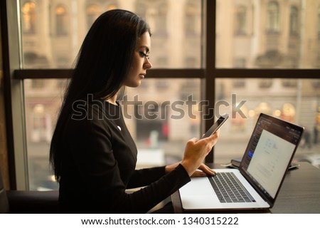 Serious confident woman leadership using apps on mobile phone during work on laptop computer, sitting in office space in evening time. Female CEO using messenger on cellphone during online job 