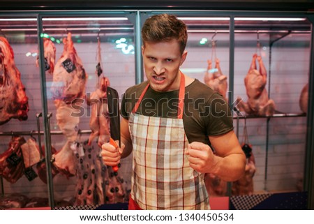 Young angry man stand in front of meet shelf in grocery store. He hold butcher knife in hand and pose. Guy maddly look on camera. Animal brawn behind.