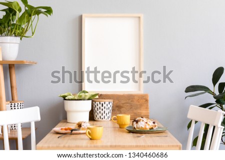 Stylish and sunny interior of kitchen space with small wooden table with mock up photo frame, design cups and tasty dessert. Scandinavian room decor with kitchen accessories and beautiful plants.