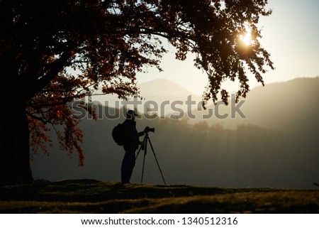 Profile of traveler tourist guy with backpack at camera tripod lit by bright setting sun in the sky standing on grassy valley on background of beautiful mountain panorama at dusk under big tree.