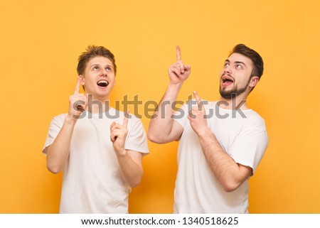 Two happy men in white T-shirts look at the place for advertising and show a finger. Portrait of a two happy young men showing thumbs up isolated over yellow background.
