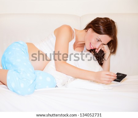 Cute pregnant woman lying down on bed at home and enjoying photo of ultrasound of her baby, happy parenthood concept