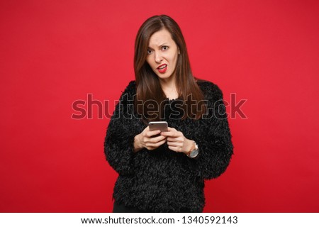 Perplexed young woman in black fur sweater using mobile phone, typing sms message isolated on bright red wall background in studio. People sincere emotions, lifestyle concept. Mock up copy space