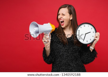Crazy young woman in black fur sweater screaming on megaphone, holding round clock isolated on bright red wall background in studio. People sincere emotions, lifestyle concept. Time is running out