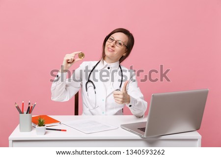 Female doctor sits at desk works on computer with medical document hold coin in hospital isolated on pastel pink background. Woman in medical gown glasses stethoscope. Healthcare medicine concept