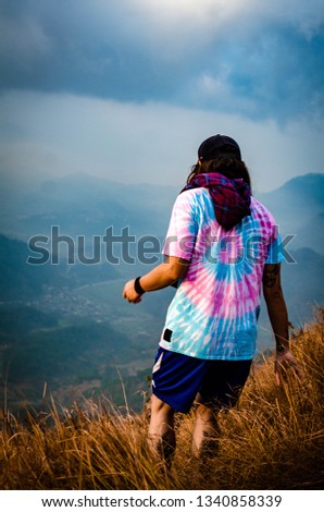 Man in a pink and blue abstract shirt walking through a field of grass while looking ahead into a range of majestic blue mountains in the distance. 