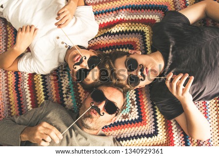 Father and two sons enjoying together lying on a colorful blanket. Three men of different ages smiling playing with fake mustache. Top view of a couple of teen and their dad with black sunglasses Royalty-Free Stock Photo #1340929361