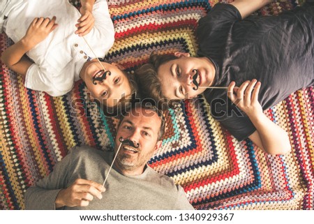 Father and two sons enjoying together lying on a colorful blanket. Three men of different ages smiling playing with fake mustache. Top view of a couple of teen and their dad. Royalty-Free Stock Photo #1340929367