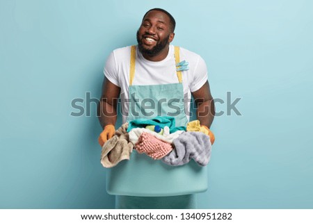 Friendly looking glad smiling dark skinned man with stubble, white teeth, holds basin with linen for washing, being in high spirit, wears casual white t shirt and apron, models over blue background