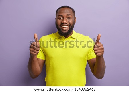 Satisfied black man gestures with both hands, shows approval gesture, keeps two thumbs raised, smiles broadly, wears yellow casual t shirt, stands against purple background. Thats nice, I like it