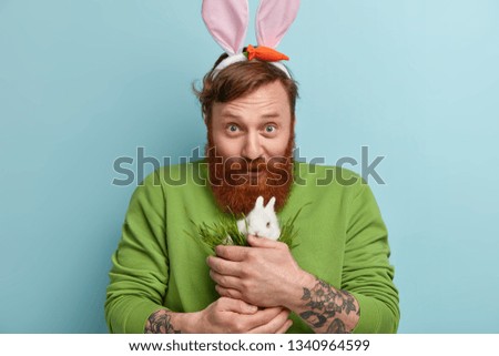Headshot of glad caring man with trendy hairstyle and thick ginger beard, buys small decorative rabbit who eats green fresh grass, carries bunny to make pleasant surprise his little daughter or son