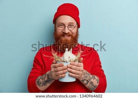 Waist up shot of friendly looking pleased hipster buys decorative white rabbit in animal shop, wants to make surprise for children, cares about pets, has tattooed arms, dressed in red clothing