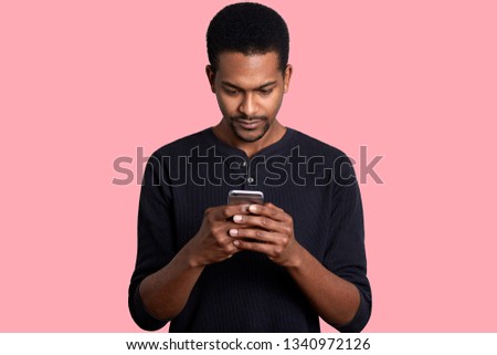 Young guy looks at the screen of smartphone with serious fecial expression. Black handsome male has important unpleasent correspondence. Boring pastime. Modern technology and Internet concept.