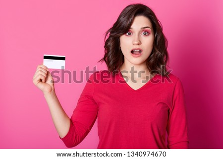 Image of beautiful shocked young woman with opened mouth isolated over rose background. Cute girl holds credit card, finds out she has no money, can not pay for purchases. Online shopping concept.