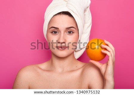 Beauty portrait of young girl with nacked shoulders. Lady holds orange, has calm facial expression. Charming female with towel wrapped around head relaxes in spa salon. Model poses in photo studio.