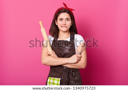 Close up portrait of thoughtful housewife with brown apron, red hair band, white t shirt. Cute female stands with crossed hands, bites her lip and can not remember right ingredients for making pie.