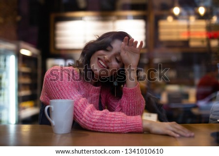 pretty armenian girl in denim dress sitting in a cafe behind a glass window with a cup of coffee and smiling