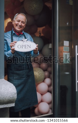 Vertical portrait of joyful old man in apron inviting customers to visit his coffee shop. He looking at camera and smiling