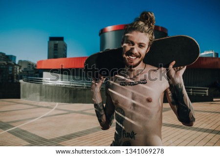 Positive smiling hipster standing in urban area while holding a skate behind his back