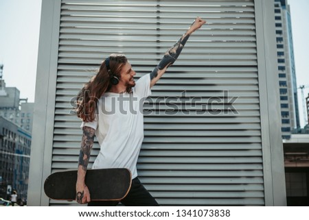 Cheerful smiling long haired man listening to music while holding a skateboard