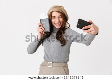 Photo of european woman 30s wearing hat taking selfie on cell phone while holding passport and travel tickets isolated over white background