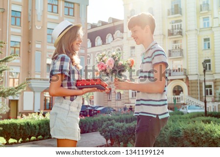 Teen boy congratulates girl with bouquet of flowers and gift, outdoor portrait couple happy youth.
