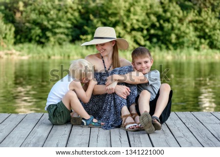 Beautiful mother with two young sons are embracing on the pier on the river bank.