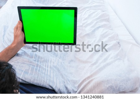 Young man lying in bed holding a greenscreen tablet.