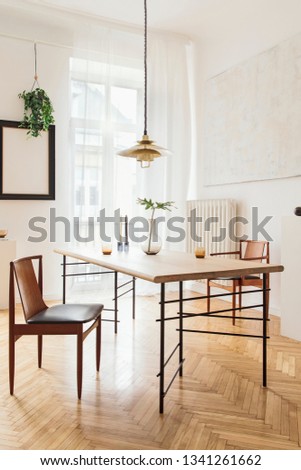 Stylish and modern dining room interior with design sharing table, chairs, gold pedant lamp, abstract paintings and elegant accessories. Tropical leafs in vase. Eclectic decor. Brown wooden parquet.