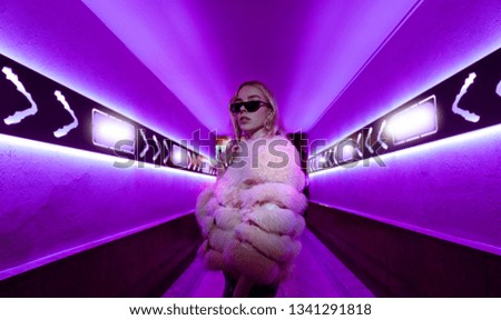 Teen hipster girl in stylish glasses and fur standing in purple neon lights elements on street wall, female teenager fashion model woman posing in city night violet bright club glow, back to 80s Royalty-Free Stock Photo #1341291818