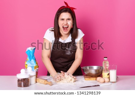 Beautiful angry cook kneads dough and screams loud, being sick and tired of preparing homemade pastry. Furious brunette woman works in kitchen and dreams of having rest. Culinary and food concept.